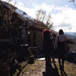 3 mensen staan buiten met een kop koffie, op de achtergrond houten huisje met rokende schoorsteen
